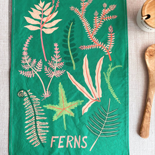 A green kitchen towel featuring a botanical illustration of various ferns in pink, green, and cream, with the word 'FERNS' printed at the bottom. The towel is styled flat-lay on a textured beige linen surface with wooden kitchen props.